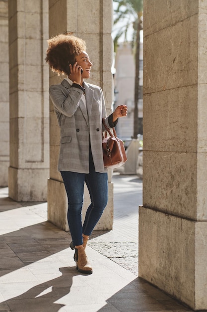 Tutta la lunghezza della donna nera soddisfatta che parla al cellulare e distoglie lo sguardo mentre si trova sul marciapiede della città sotto il sole