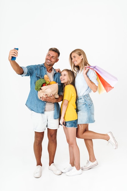 Tutta la lunghezza di una famiglia allegra che indossa abiti casual in piedi isolata su bianco, facendo la spesa insieme, portando borse, facendo un selfie