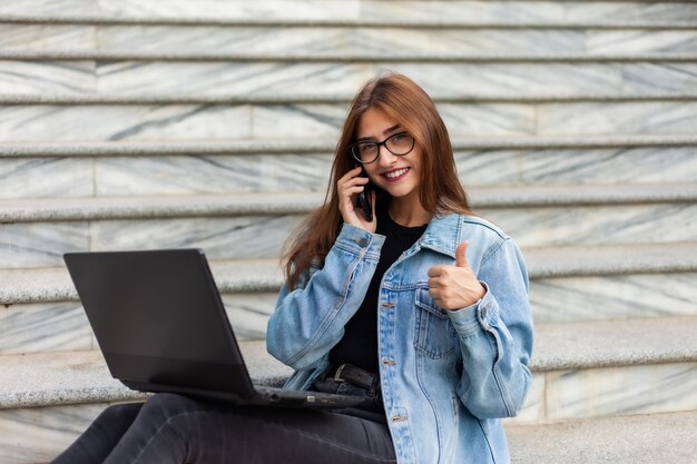 Tutti in affari. La giovane donna alla moda in una giacca di jeans e occhiali usa un computer portatile e parla al telefono mentre era seduto sulle scale in città. Lavoro a distanza.