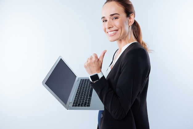 È tutto bellissimo. Donna positiva allegra felice che sorride e che mostra segno GIUSTO mentre tiene un computer portatile