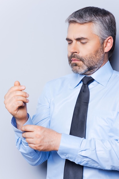 Tutto dovrebbe essere perfetto. Fiducioso uomo maturo in camicia e cravatta che si aggiusta la manica mentre sta in piedi su uno sfondo grigio