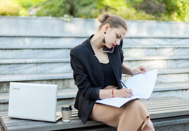 Tutto in una donna d'affari che parla al telefono scrive nella cartella con la carta seduta su una panchina con un computer portatile all'aperto