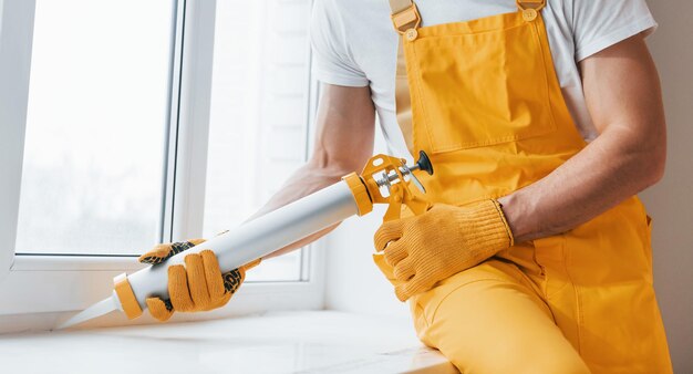 Tuttofare in uniforme gialla lavora con colla per finestre all'interno Concezione di ristrutturazione della casa