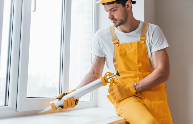 Tuttofare in uniforme gialla lavora con colla per finestre all'interno Concezione di ristrutturazione della casa