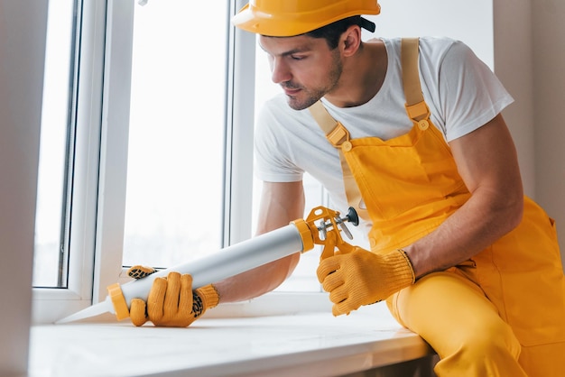 Tuttofare in uniforme gialla lavora con colla per finestre all'interno Concezione di ristrutturazione della casa