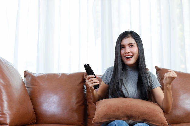 TV e concetto di felicità. Bella donna asiatica in casual seduta sul divano in soggiorno, tenendo il telecomando della televisione e guardando qualcosa con una faccia sorridente felice. Concetto di stile di vita.