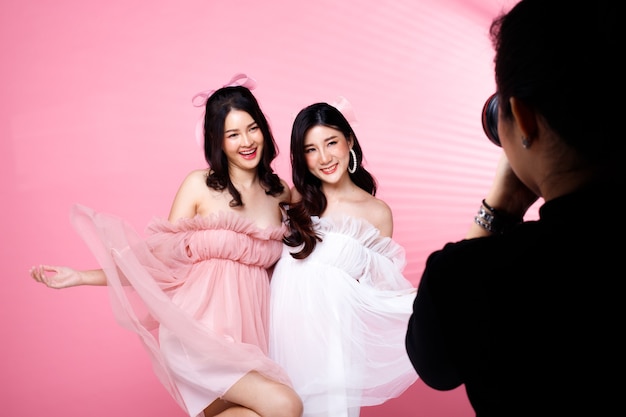 Two Fashion Beauty Women ha i capelli neri che saltano ed esprimono un sorriso. Le ragazze asiatiche indossano un abito rosa su un muro di tonalità rosa con muro di pomeriggio ombra striscia di finestra, copia spazio