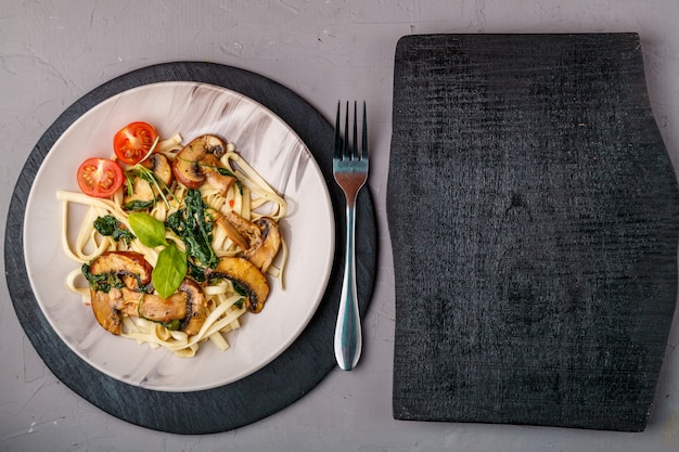 Udon con funghi e spinaci in un piatto su una superficie di cemento vicino a una forchetta e una tavola di legno