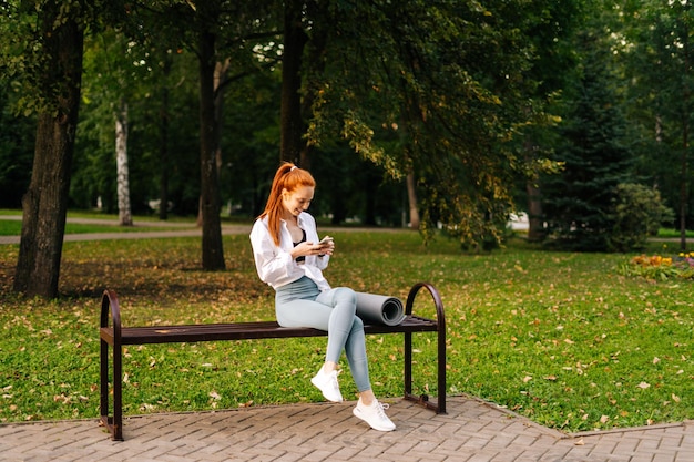 UFA RUSSIA 7 SETTEMBRE 2020 Giovane donna gioiosa dai capelli rossi che indossa abbigliamento sportivo guardando video su smartphone seduto panchina al parco cittadino Donna attraente che chiacchiera con gli amici utilizzando il cellulare