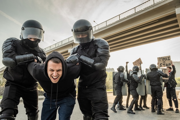 Ufficiale antisommossa in caschi che torce le braccia di un manifestante aggressivo in un maglione con cappuccio mentre lo arresta per strada