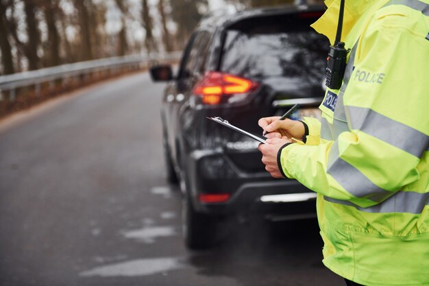 Ufficiale di polizia maschio in uniforme verde che sta con il blocco note vicino all'automobile.