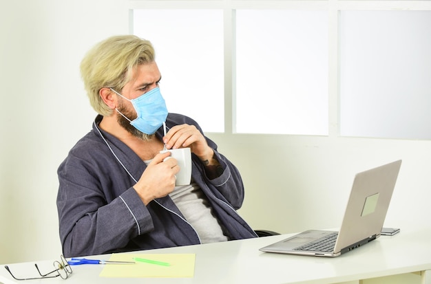 Ufficio da casa Lavoro a distanza Lavoratore in maschera medica Tutto ciò di cui hai bisogno per una giornata produttiva Totalmente protetto Indossare la maschera protegge dal coronavirus L'uomo in maschera beve tè caffè usando la cannuccia Igiene seria