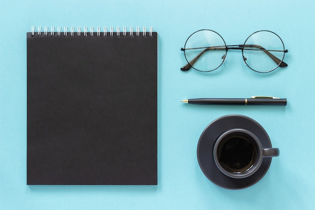 Ufficio o posto di lavoro a casa. Blocco note di colore nero, tazza di caffè, occhiali, penna su sfondo blu