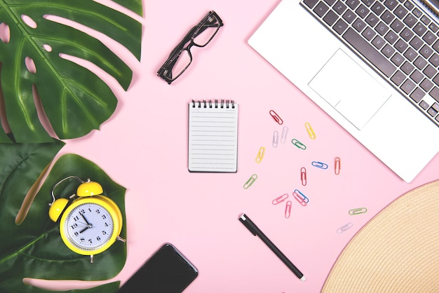 Ufficio sul posto di lavoro con computer portatile agenda giornaliera telefono penna e occhiali con decorazione tropicale Vacanze di lavoro piatto per i social media su sfondo rosa