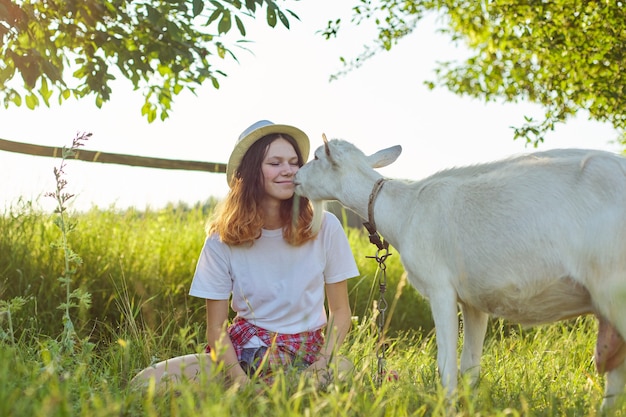 Umorismo, capra bianca dell'azienda agricola che bacia ragazza dell'adolescente. Paesaggio tramonto panoramico estivo