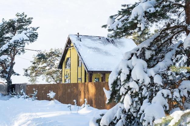 Un'accogliente casa gialla nella neve in inverno nel villaggio è circondata da pini Tetto innevato riscaldamento e tubi di ventilazione finestre trapezoidali