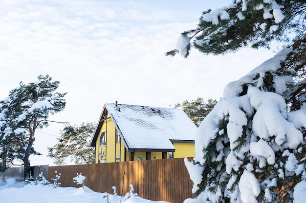 Un'accogliente casa gialla nella neve in inverno nel villaggio è circondata da pini Tetto innevato riscaldamento e tubi di ventilazione finestre trapezoidali