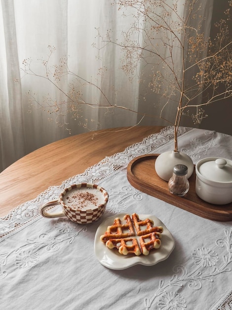 Un'accogliente pausa caffè, cappuccino e waffle con latte condensato su un tavolo rotondo di legno.