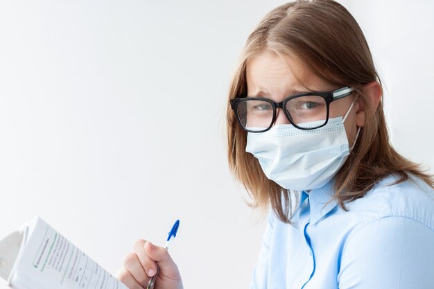 Un'adolescente, bionda, con una camicia blu, una maschera medica e occhiali su uno sfondo bianco guarda la telecamera, con in mano una penna e un taccuino.