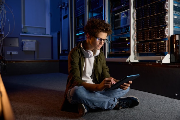 Un adolescente che lavora nell'informatica è seduto sul pavimento di una moderna sala server e sta lavorando su un tablet