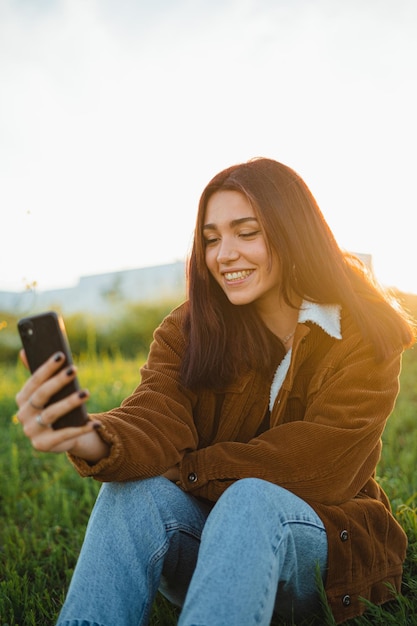 Un adolescente che videochiama un amico seduto su un prato verde durante il tramonto in primavera