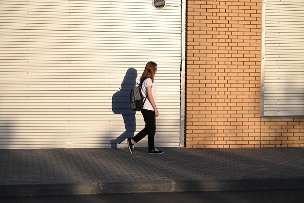 Un'adolescente con pantaloni neri e maglietta bianca cammina per strada con uno zaino nero sulla schiena. Ritorno a scuola