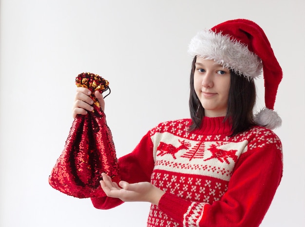 Un'adolescente con un cappello da Babbo Natale e un maglione rosso di Natale tiene in mano una borsa rossa con un regalo. Il concetto di Natale e Capodanno