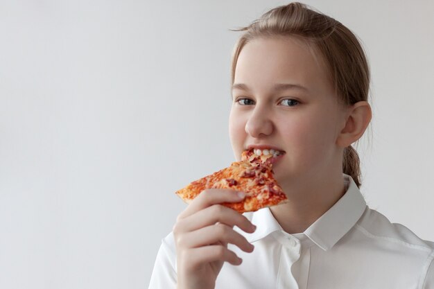 Un'adolescente con una camicia bianca si mette in bocca una fetta di pizza e dà un morso, guarda la telecamera.