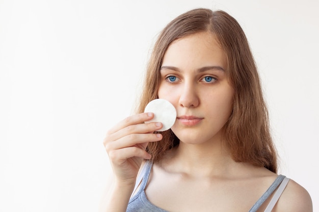 Un'adolescente di quindici anni con lunghi capelli castani, occhi azzurri e pelle pulita si strofina il viso con un batuffolo di cotone. Cura di sé, cura della pelle. Copia spazio.