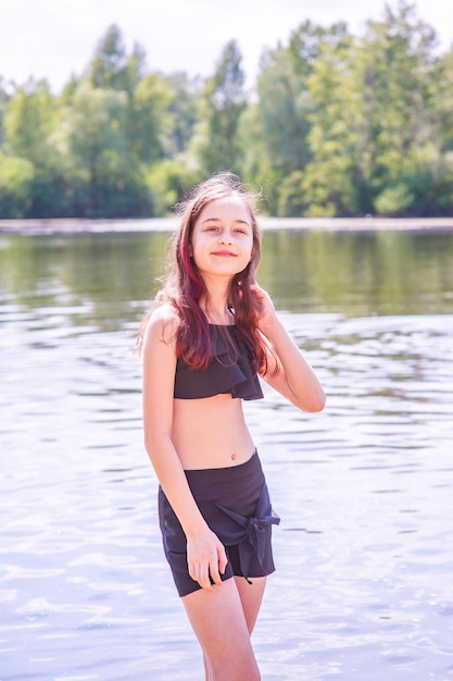 Un'adolescente in riva al fiume in costume da bagno nero e pantaloncini neri in estate. Bambini e riposo.