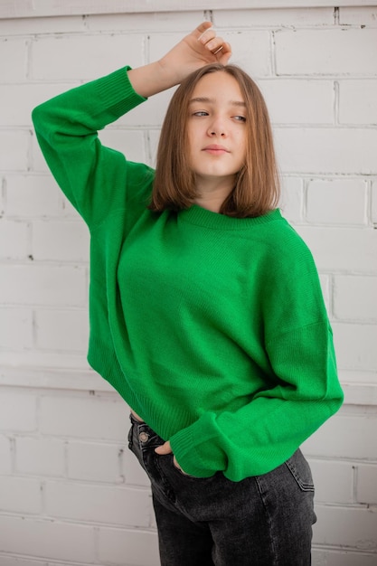Un'adolescente snella in jeans e un maglione verde posa contro un muro di mattoni bianchi. bellezza naturale, nessun trucco, illuminazione naturale. riprese di moda. spazio per il testo. Foto di alta qualità