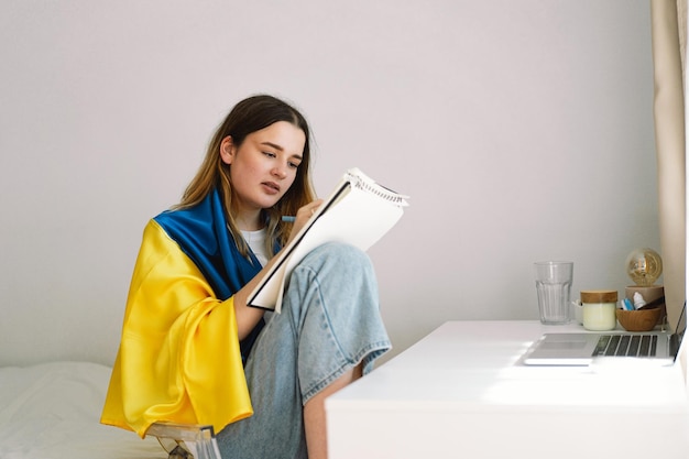 Un'adolescente ucraina è avvolta nella bandiera ucraina e sta disegnando o facendo i compiti in camera da letto Un adolescente studia a casa Il concetto di studi durante la guerra in Ucraina