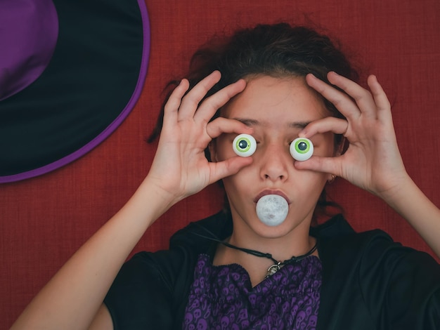 Un'adolescente vestita da strega si copre gli occhi di caramelle e soffia una pallina di gomma da masticare