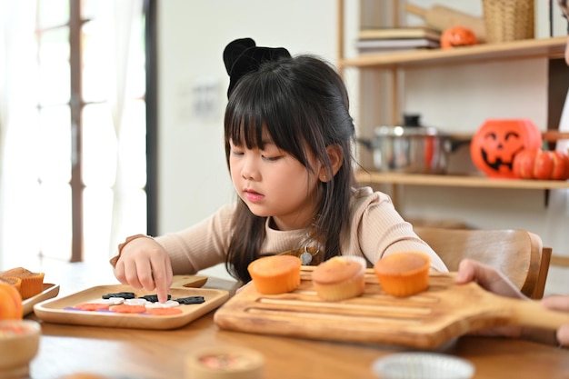 Un'adorabile bambina asiatica che si concentra sulla preparazione e sulla decorazione dei cupcakes di Halloween