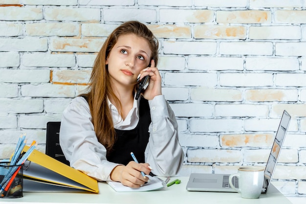 Un adorabile studente seduto dietro la scrivania e parlando al telefono e prendendo appunti Foto di alta qualità