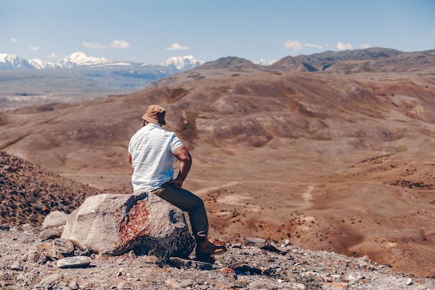 Un adulto brutale maschio barbuto con un cappello panama e una camicia bianca siede su una roccia e guarda un favoloso paesaggio di montagna con un canyon. Vista naturale dall'alto.