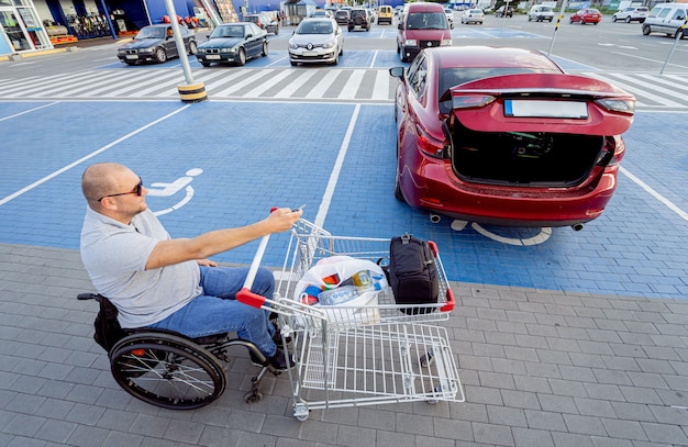 Un adulto disabile su sedia a rotelle mette gli acquisti nel bagagliaio di un'auto nel parcheggio di un supermercato