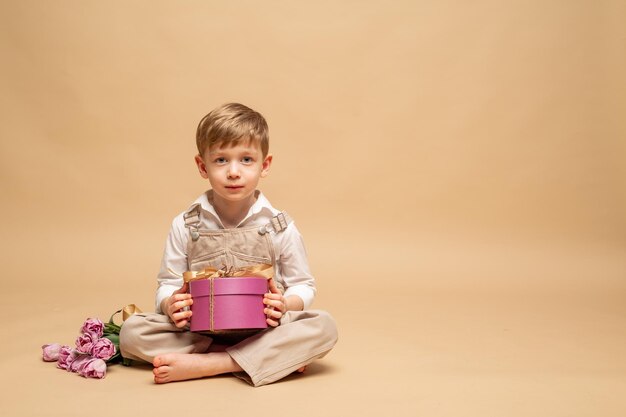 un affascinante bambino biondo di quattro anni con una tuta beige e una camicia bianca si siede e tiene in mano un regalo lilla