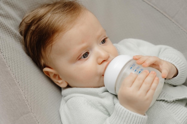 Un affascinante bambino di 5 mesi dagli occhi azzurri giace a letto e beve il latte da un biberon con un biberon