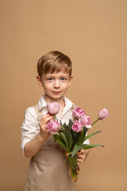 un affascinante bambino di quattro anni in tuta beige e camicia bianca tiene in mano un mazzo di tulipani rosa