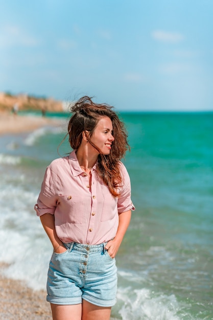 Un'affascinante giovane donna con i capelli lunghi e con indosso una maglietta cammina lungo la spiaggia in una giornata di sole