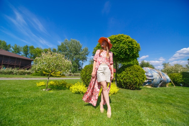 Un'affascinante ragazza con un leggero prendisole estivo e un pareo sta camminando in un parco verde. Gode di calde giornate estive soleggiate.