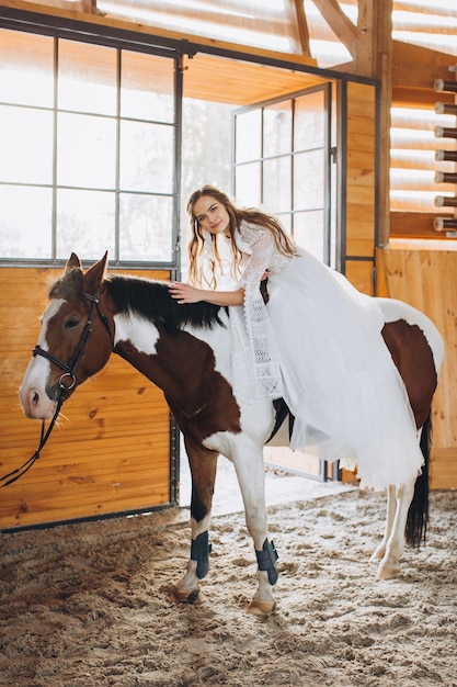 Un'affascinante sposa boho cavalca un cavallo in un ranch al tramonto in inverno