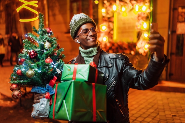 Un afroamericano sorridente con un albero di Natale e regali in mano prende un selfie per strada