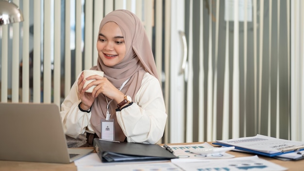 Un'allegra donna d'affari asiatica-musulmana sta prendendo il caffè mentre lavora al suo portatile alla scrivania