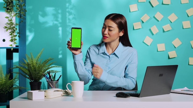 Un'allegra impiegata asiatica in possesso di uno smartphone con schermo verde a chiave cromatica, che presenta informazioni. Segretaria professionale in un luogo di lavoro rilassato e colorato sullo sfondo blu dello studio