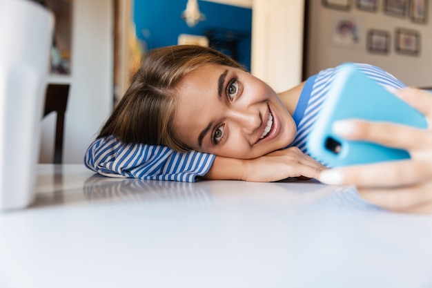 un allegro ottimista sorridente giovane ragazza carina a casa al tavolo utilizzando il telefono cellulare.