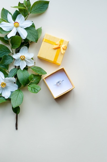 Un anello con una grossa pietra in una scatola gialla accanto a un ramo con fiori di narciso su fondo beige