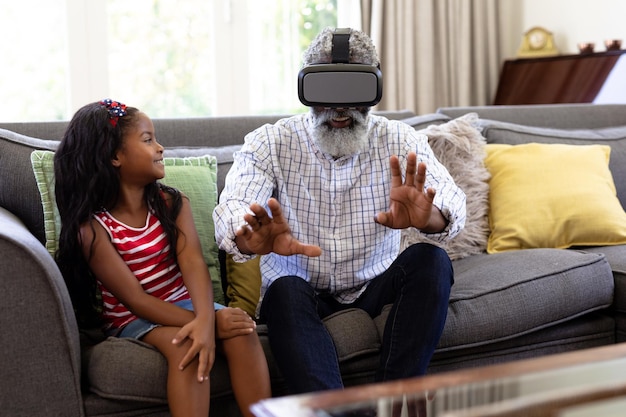 Un anziano afroamericano e sua nipote si godono il tempo a casa insieme, seduti su un divano, indossando un visore VR