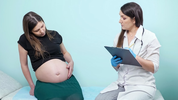 Un'attenta dottoressa in uniforme con appunti tocca la pancia della donna incinta seduta sul divano nell'ufficio della clinica contemporanea all'appuntamento